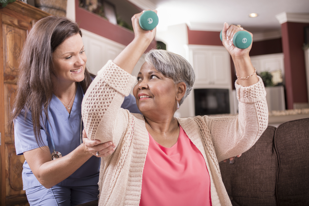 Caring, Latin descent home healthcare nurse conducts physical therapy exercises with African descent senior adult patient at home, assisted living, or nursing home setting.  She helps her with arm strengthening exercises using dumbbells.